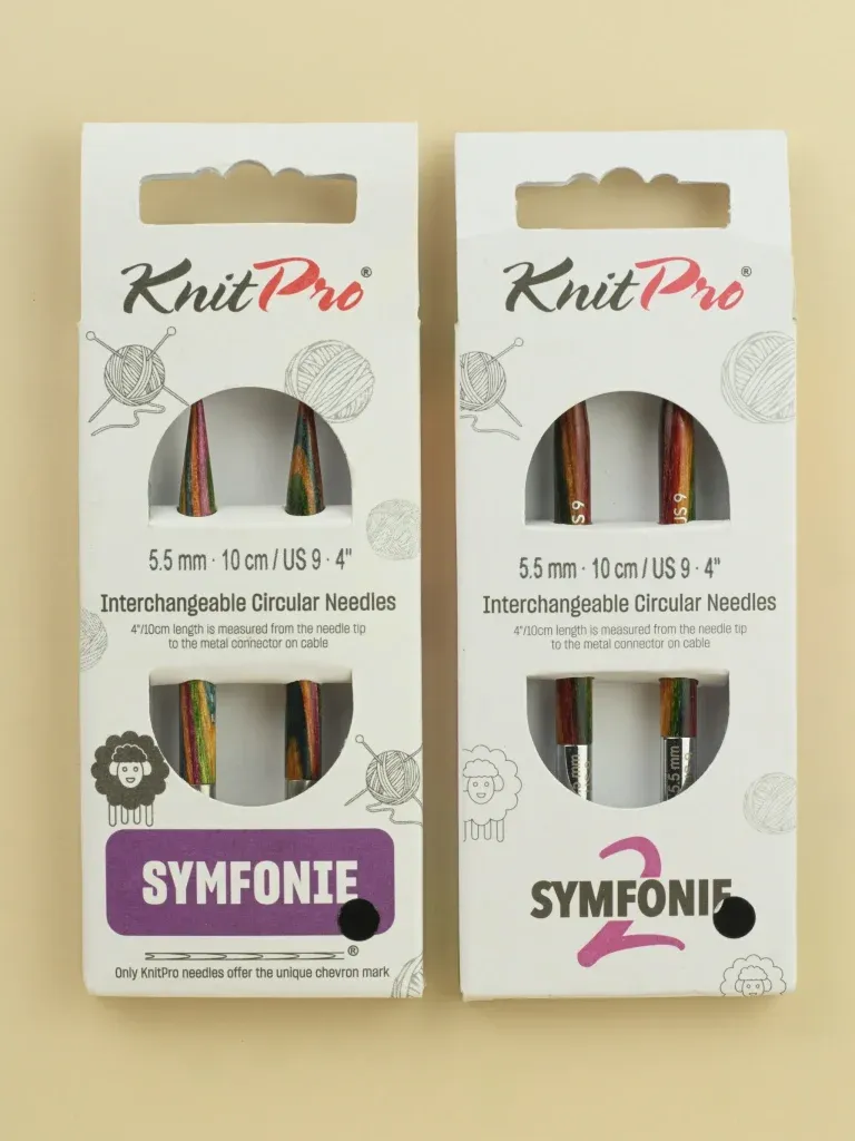 Two small white cardboard packaging boxes lie side by side. At first glance they look very similar. Both say KnitPro 5.5mm, 10cm/4" interchangeable circular needles. The white cardboard has small black-and-white line drawings of balls of wool and sheep on it. There are circular windows that show colourful knitting needles inside. The pack on the left says Symfonie, while the pack on the right says Symfonie 2.
