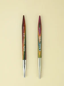 Two interchangeable circular knitting needles lie side by side, points facing to the top of the image. They look very, very similar. Both are made from wood with multi-coloured surfaces. The one on the left is slightly glossier and the colours seem a little darker and richer. It has the words KnitPro 5.5mm US9 printed on the body. The one on the right is a little paler and has less of a sheen. It has 5.5mm US9 printed on it. Both end in silver, tapered cable connectors. The one on the left is shorter and shinier, and also has 5.5mm printed on it.