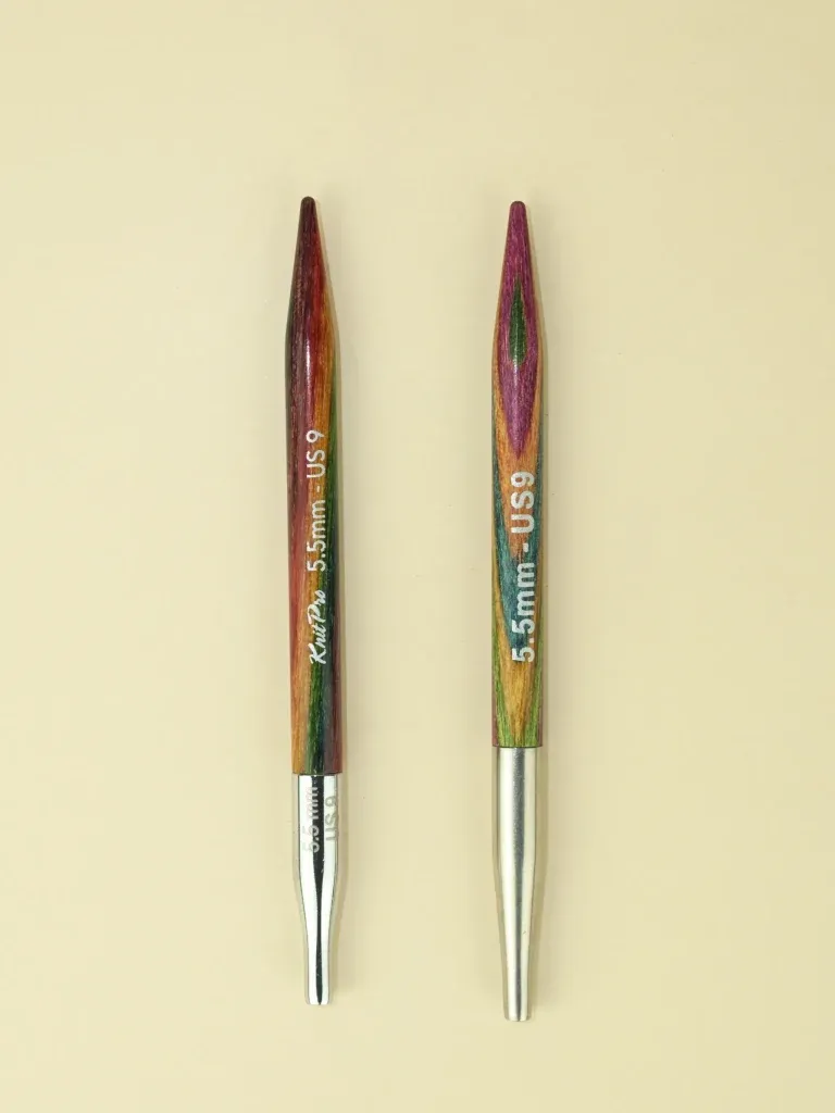 Two interchangeable circular knitting needles lie side by side, points facing to the top of the image. They look very, very similar. Both are made from wood with multi-coloured surfaces. The one on the left is slightly glossier and the colours seem a little darker and richer. It has the words KnitPro 5.5mm US9 printed on the body. The one on the right is a little paler and has less of a sheen. It has 5.5mm US9 printed on it. Both end in silver, tapered cable connectors. The one on the left is shorter and shinier, and also has 5.5mm printed on it.
