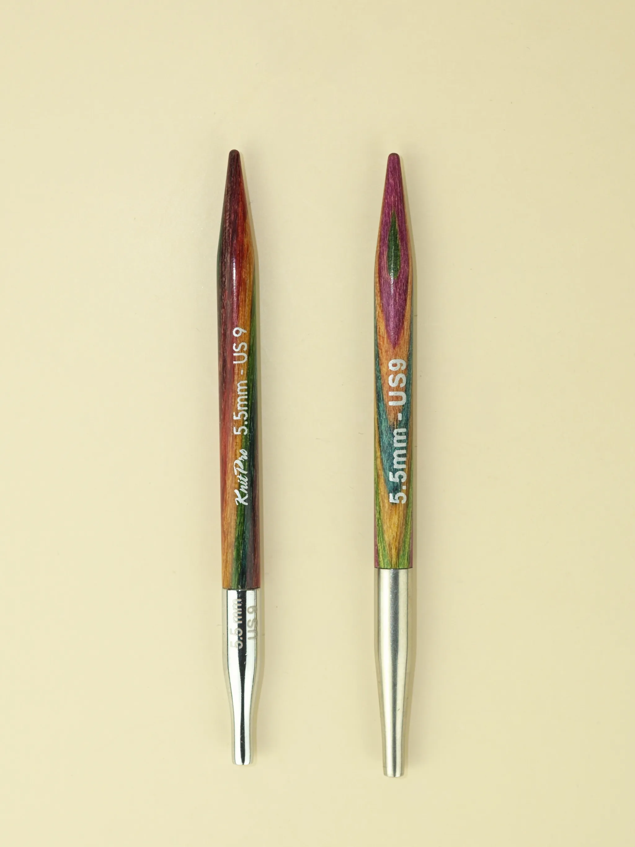 Two interchangeable circular knitting needles lie side by side, points facing to the top of the image. They look very, very similar. Both are made from wood with multi-coloured surfaces. The one on the left is slightly glossier and the colours seem a little darker and richer. It has the words KnitPro 5.5mm US9 printed on the body. The one on the right is a little paler and has less of a sheen. It has 5.5mm US9 printed on it. Both end in silver, tapered cable connectors. The one on the left is shorter and shinier, and also has 5.5mm printed on it.